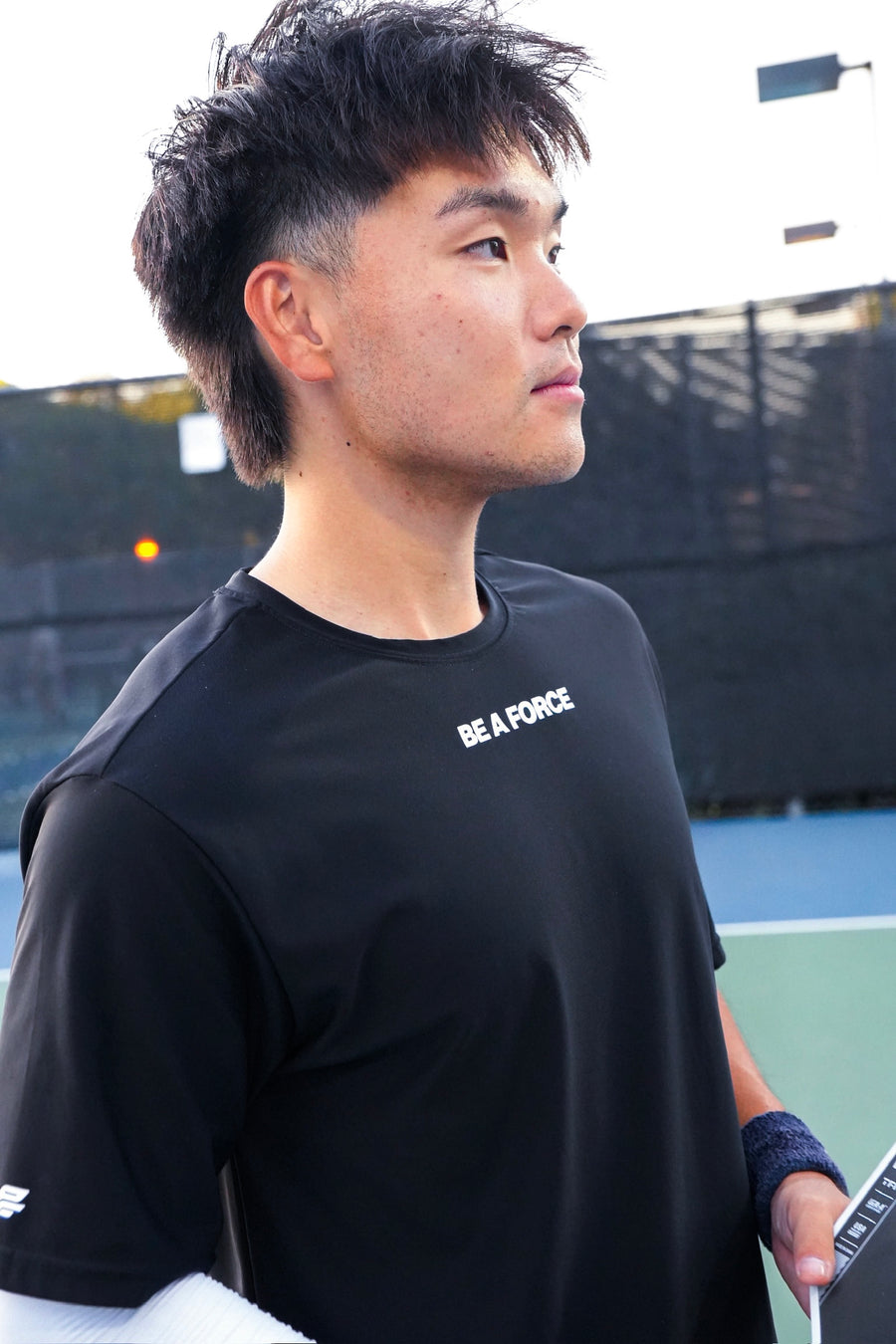 Person wearing a black t-shirt with 'BE A FORCE' text on a tennis court, black