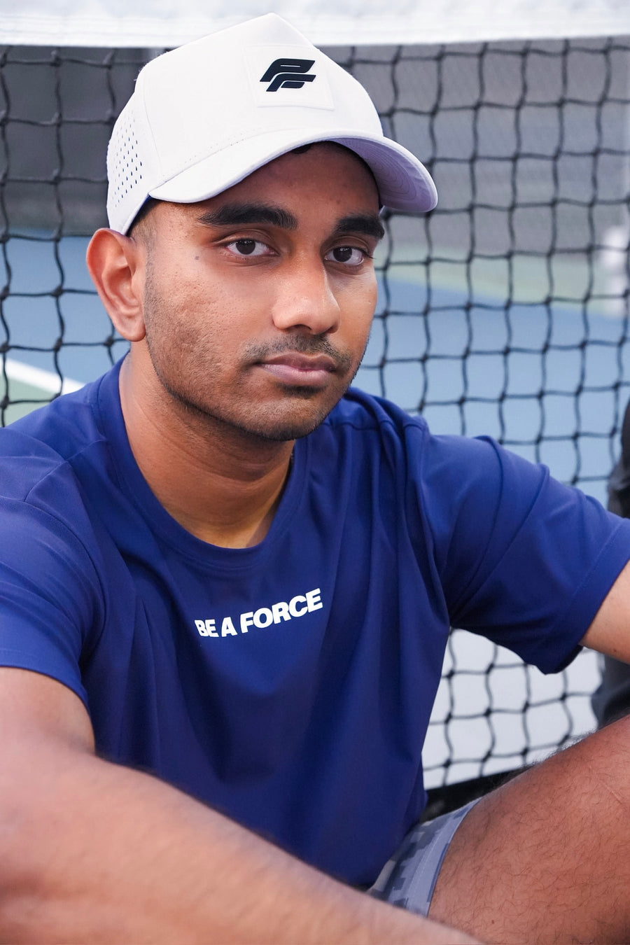 Man wearing a blue shirt with 'Be A Force' text and a white cap, sitting in front of a tennis net, navy