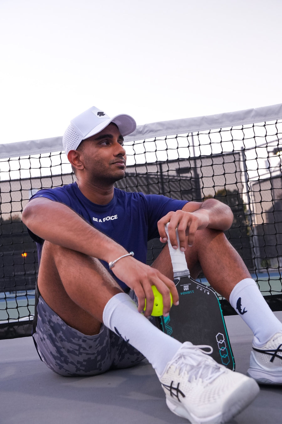 Man sitting on a tennis court with a tennis ball and a skateboard, wearing a blue shirt and white cap, navy