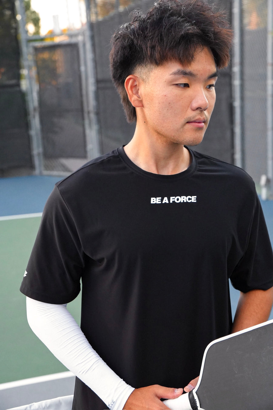Person wearing a black t-shirt with 'BE A FORCE' text on a tennis court, black