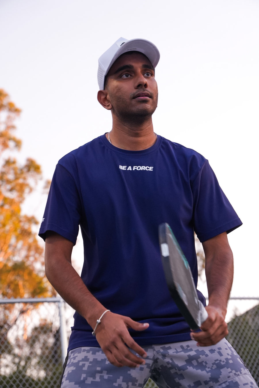 Man in a blue t-shirt and cap holding a paddle on a tennis court, navy
